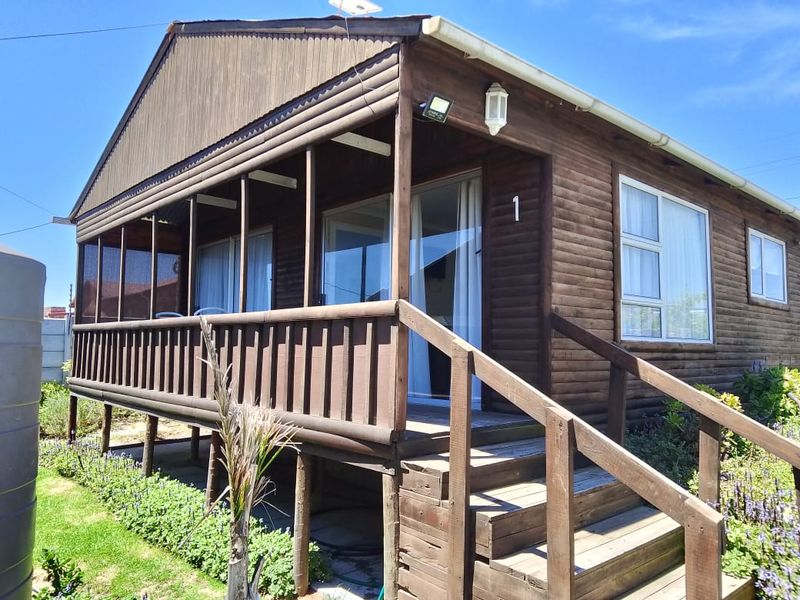 A timber-clad cabin featuring a raised covered veranda, wooden staircase, and sliding glass doors, set within a landscaped garden area.