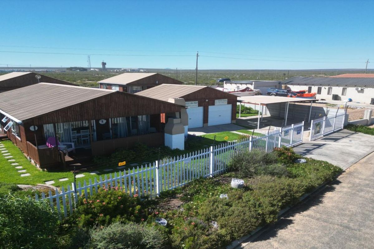 Aerial view of a coastal property featuring timber-clad buildings, a double garage, carport, and a well-maintained garden with picket fencing.