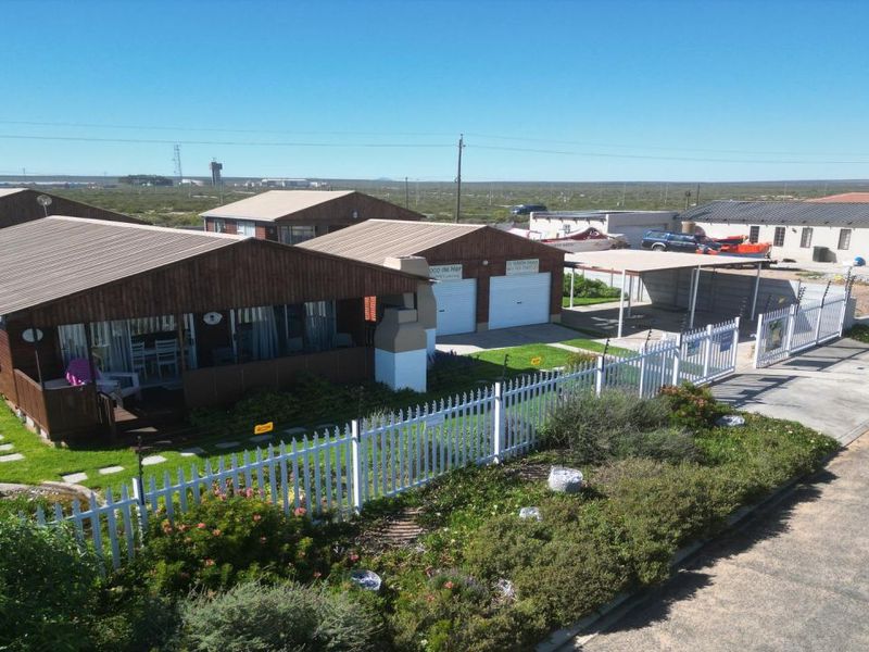Aerial view of a coastal property featuring timber-clad buildings, a double garage, carport, and a well-maintained garden with picket fencing.