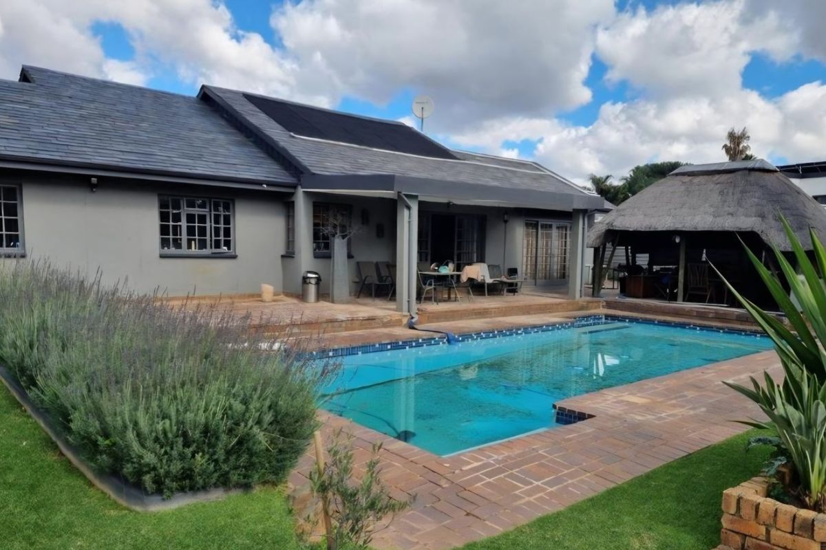 Rear exterior view featuring a swimming pool, paved deck, covered patio area, and a traditional thatched lapa, set against a well-maintained garden.