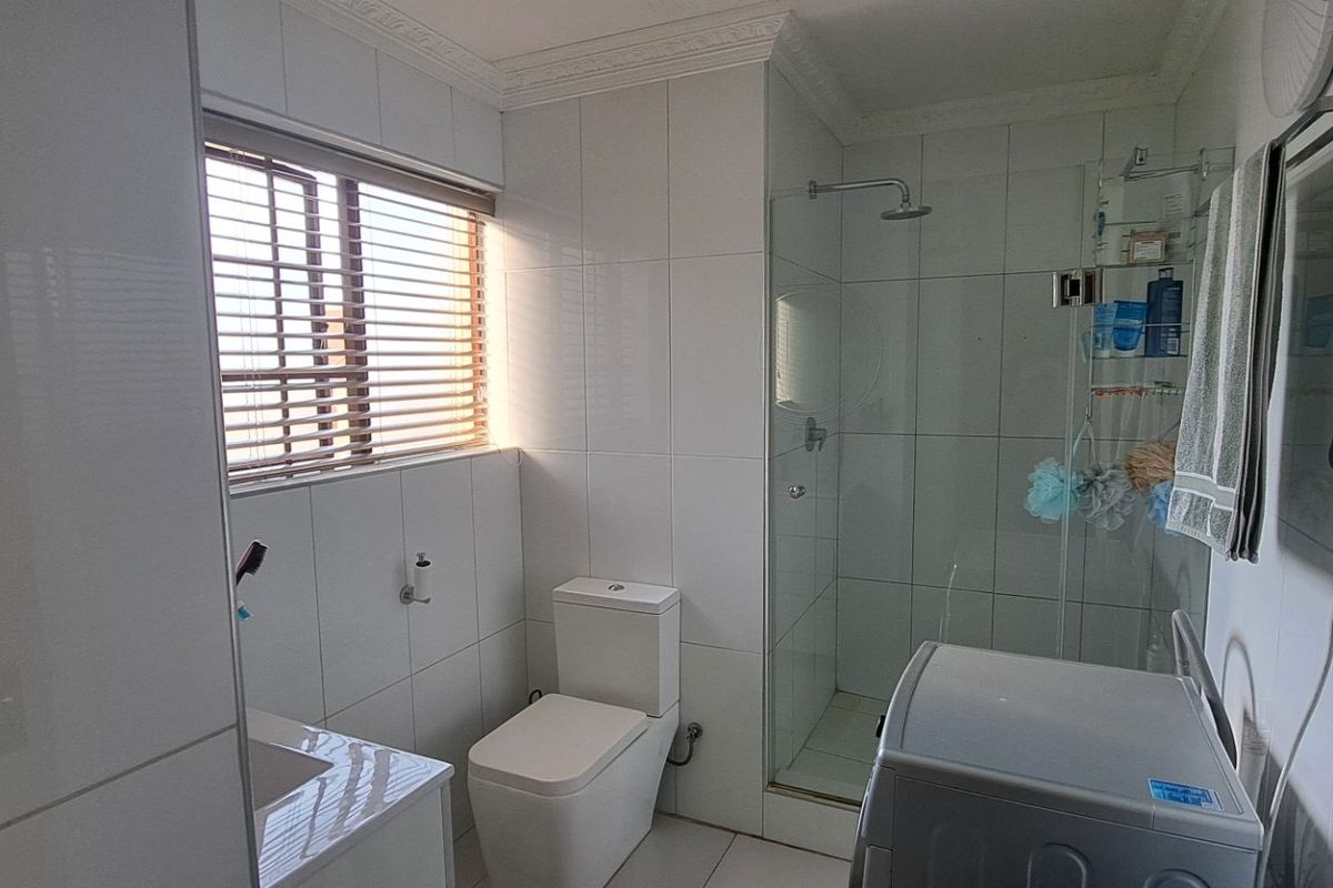 A clean, modern bathroom featuring white wall tiles, a glass-enclosed shower, a vanity basin, and a toilet, with natural light from a window.