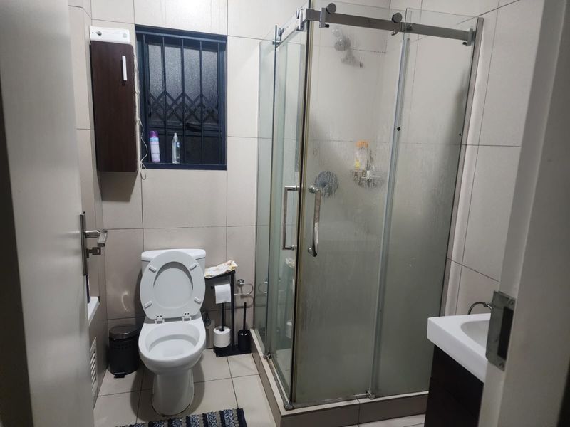 A functional bathroom featuring a glass-enclosed shower, wall-mounted vanity, and tiled finishes, complete with a window fitted with security bars.