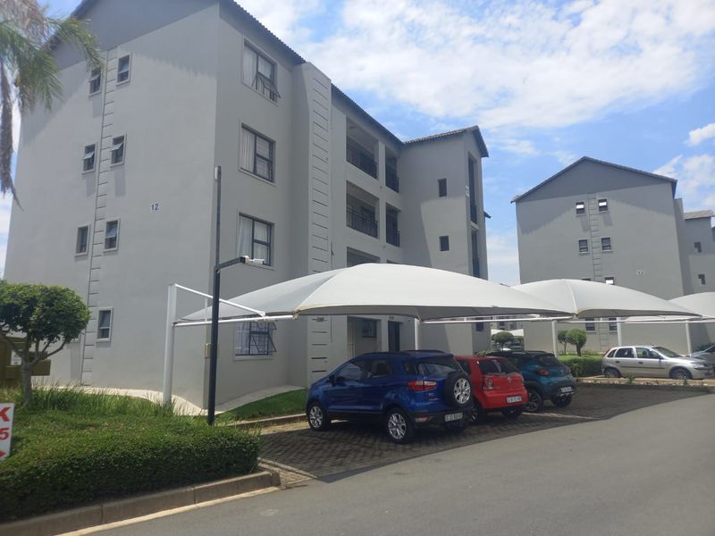 Exterior view of a modern multi-story apartment complex featuring paved parking bays with shade cloth carports and well-maintained building facades.