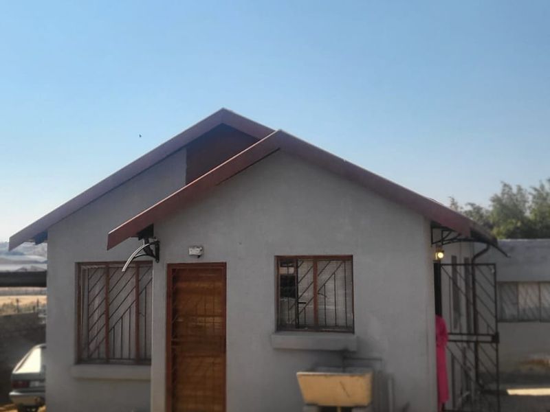 A modest single-story residential dwelling featuring a pitched roof, security bars on windows, and an external concrete wash basin on a paved lot.