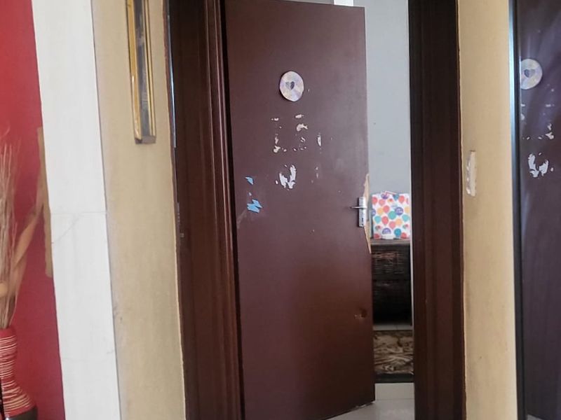 A narrow residential hallway featuring tiled flooring and recessed lighting, leading to an adjacent room with a damaged interior door.