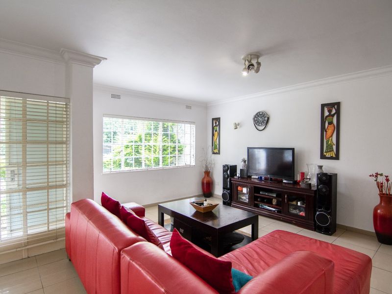 A bright and spacious living room featuring neutral tiled flooring, white walls, and large windows fitted with venetian blinds.