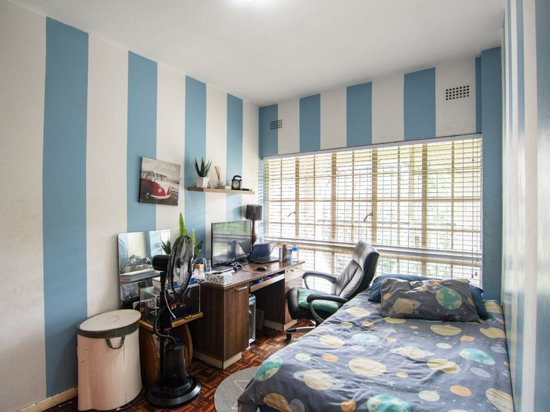 A bedroom featuring parquet flooring and large windows with venetian blinds, currently utilized as a combined sleeping and workspace area.