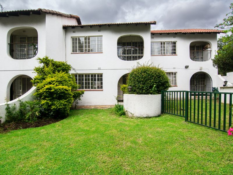 Rear view of a multi-story apartment building featuring white textured walls, tiled roofing, and a private garden area with a metal fence.