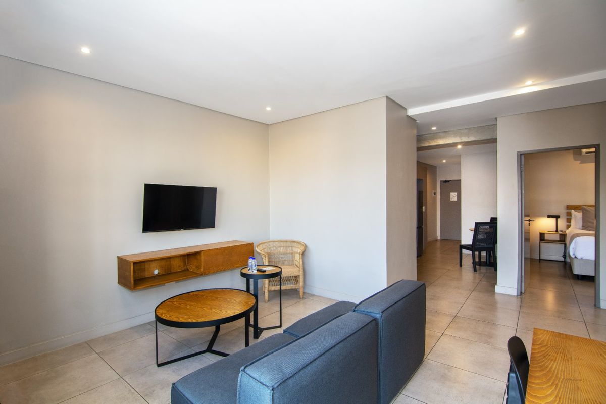 A modern living room featuring neutral walls, tiled flooring, recessed downlighting, and a wall-mounted shelving unit, leading into a hallway and bedroom area.