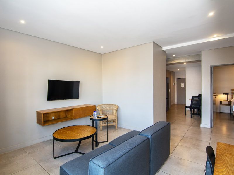 A modern living room featuring neutral walls, tiled flooring, recessed downlighting, and a wall-mounted shelving unit, leading into a hallway and bedroom area.