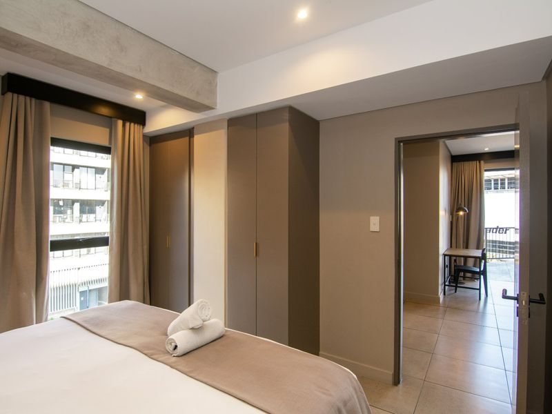 Modern bedroom featuring built-in cupboards, tiled flooring, and an exposed concrete beam, with an adjoining room visible through the doorway.
