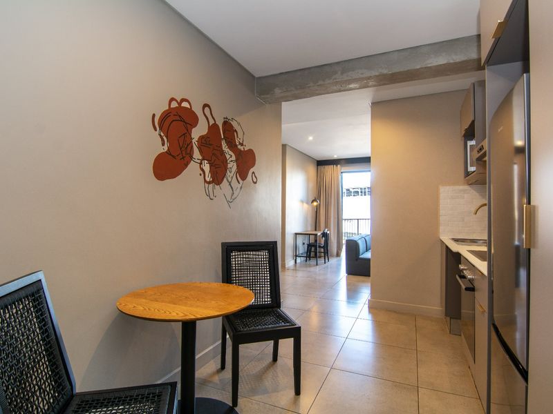 Modern open-plan apartment featuring a compact kitchen with integrated appliances, tiled flooring, and an exposed concrete beam, leading to a bright living area.