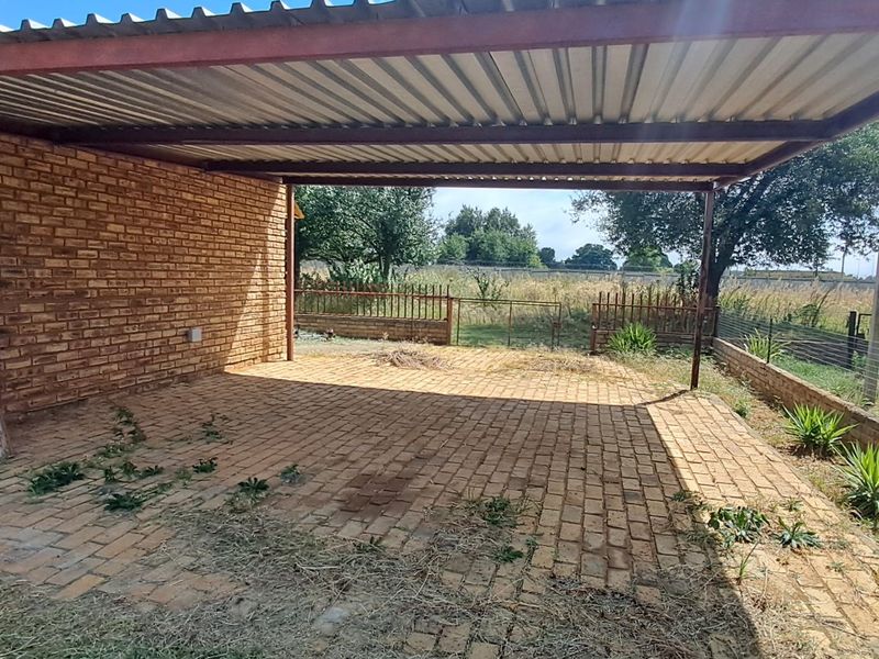 A covered outdoor patio area featuring a corrugated metal roof, steel support structure, and brick paving, overlooking a fenced garden space.