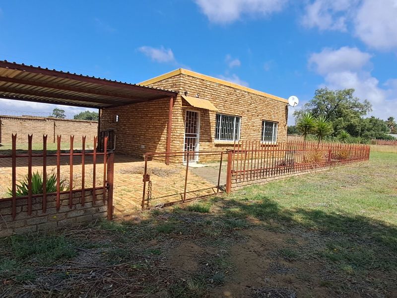 A detached face brick house featuring a carport, security fencing, and a large surrounding yard area.