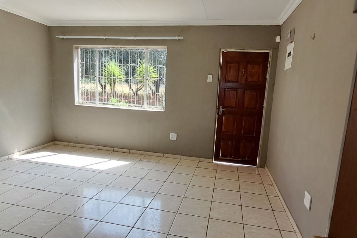 An empty living room featuring tiled flooring, a timber entrance door, and security bars on the window, presenting a blank canvas for renovation.