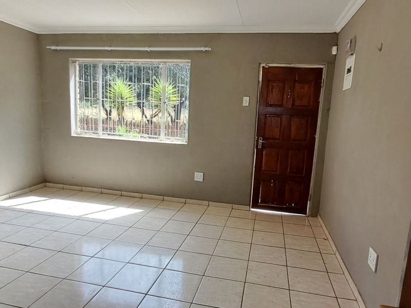 An empty living room featuring tiled flooring, a timber entrance door, and security bars on the window, presenting a blank canvas for renovation.