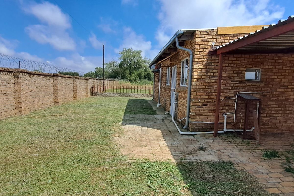Rear exterior view showing a face brick house with a paved patio area, carport, and high perimeter wall with security razor wire.