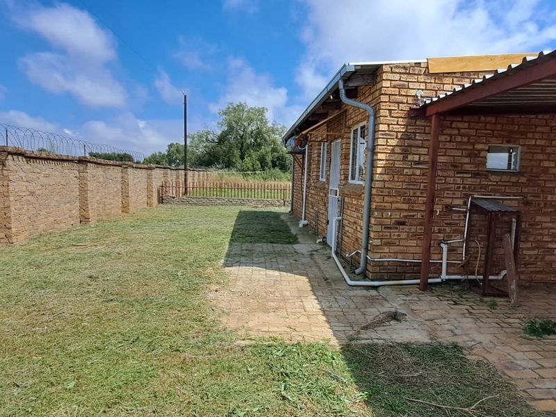 Rear exterior view showing a face brick house with a paved patio area, carport, and high perimeter wall with security razor wire.