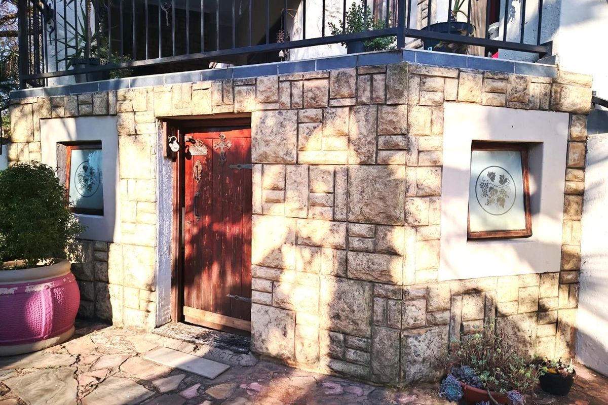 A rear exterior view featuring stone-clad walls, a timber entrance door, frosted glass windows, and a wrought iron balcony railing above a paved patio area.