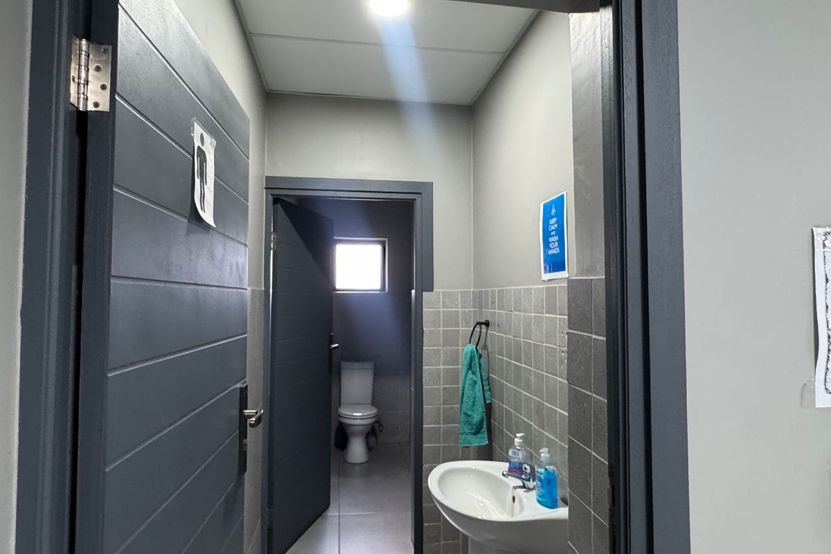 A functional industrial-style bathroom featuring a pedestal sink, tiled wall finishes, and a separate toilet cubicle with natural light.