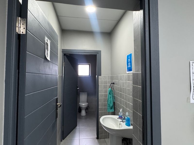 A functional industrial-style bathroom featuring a pedestal sink, tiled wall finishes, and a separate toilet cubicle with natural light.