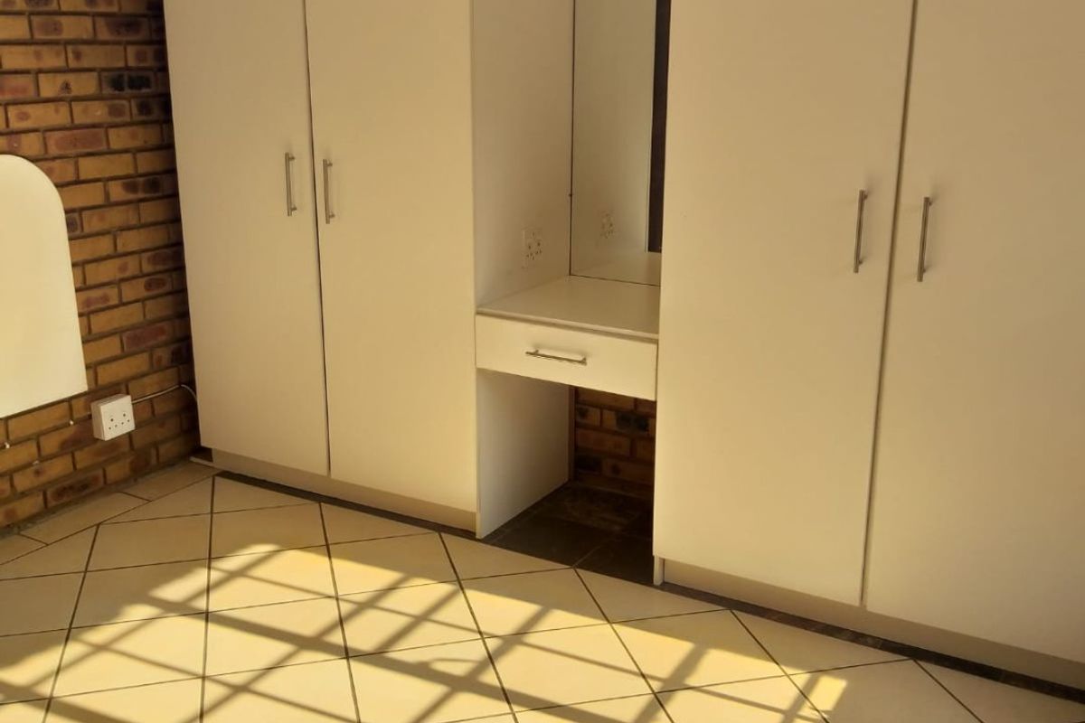 A bedroom featuring built-in white cupboards with an integrated vanity unit, tiled flooring, and an exposed brick feature wall.