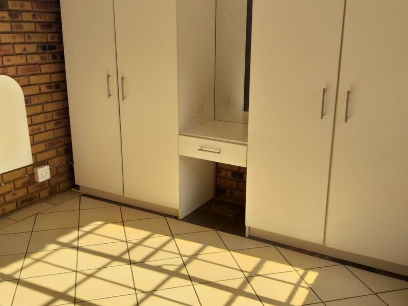 A bedroom featuring built-in white cupboards with an integrated vanity unit, tiled flooring, and an exposed brick feature wall.