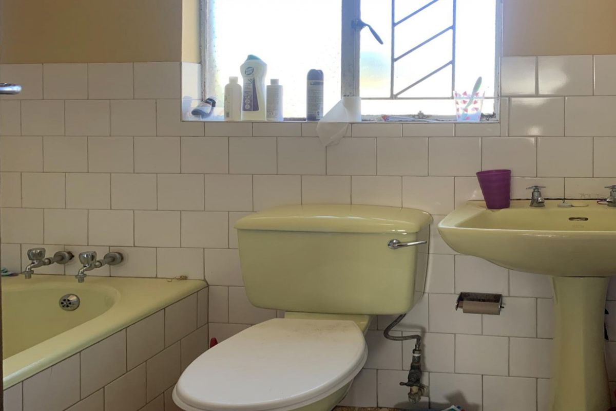 A dated bathroom featuring a pastel-colored pedestal basin, matching bathtub, and toilet, finished with white wall tiles and a window with security bars.