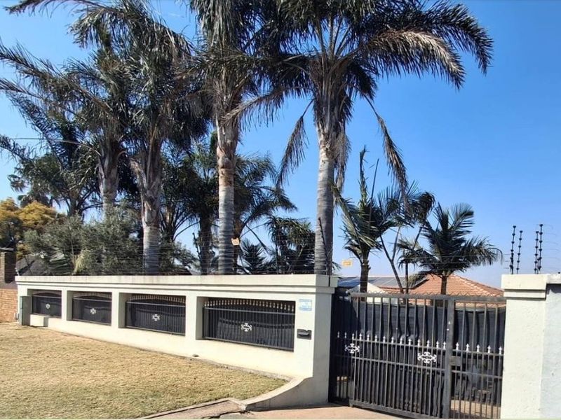 Front view of the property featuring a secure perimeter wall with electric fencing and a wrought iron entrance gate, surrounded by mature palm trees.