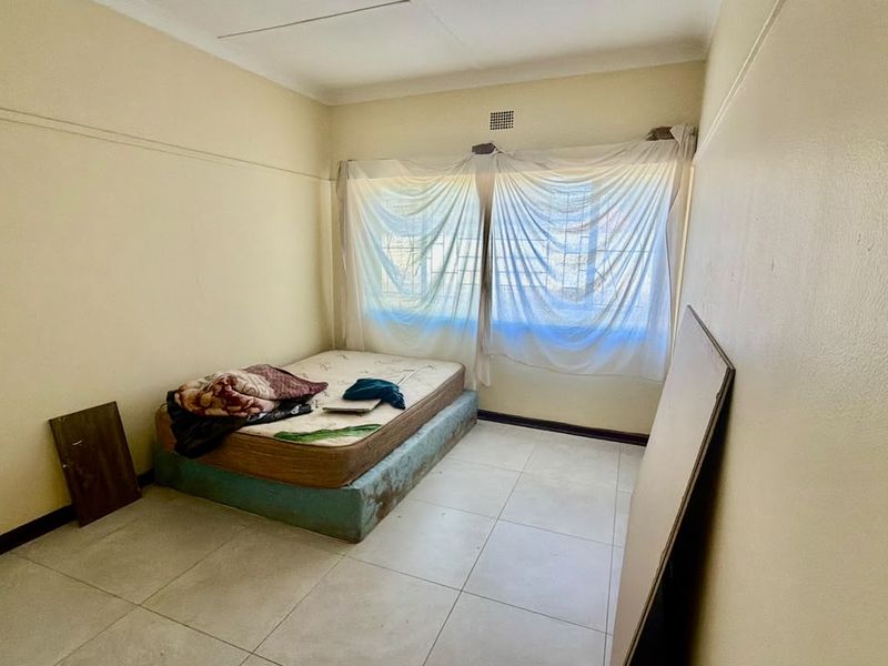 A simple bedroom featuring tiled flooring, security bars on the windows, and neutral wall finishes, offering a blank canvas for potential renovation.