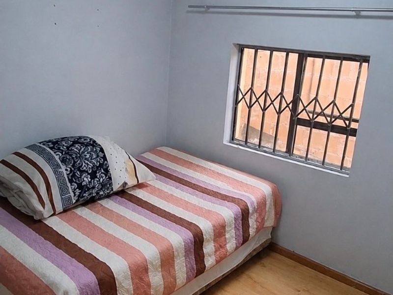 A compact bedroom featuring light-toned laminate flooring, a window with a security gate, and a simple ceiling light fixture.