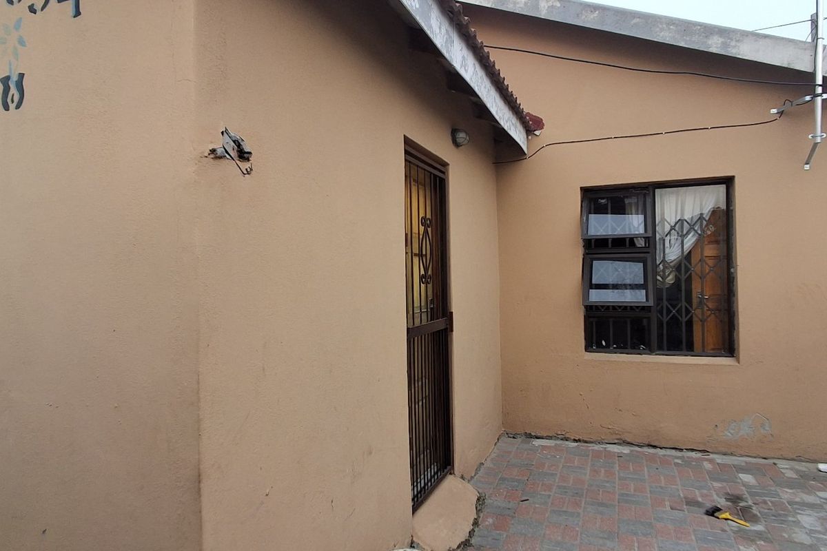 Rear exterior view of the house featuring paved patio area, security gates, and aluminium window frames, showing signs of wear and maintenance requirements.