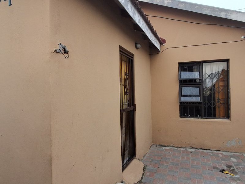 Rear exterior view of the house featuring paved patio area, security gates, and aluminium window frames, showing signs of wear and maintenance requirements.
