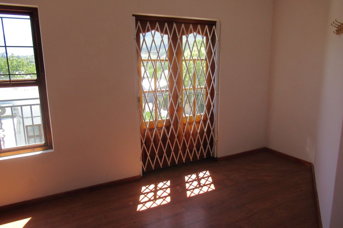 An empty bedroom featuring laminate flooring, timber-framed windows, and double doors with a security gate, offering natural light and a neutral interior.