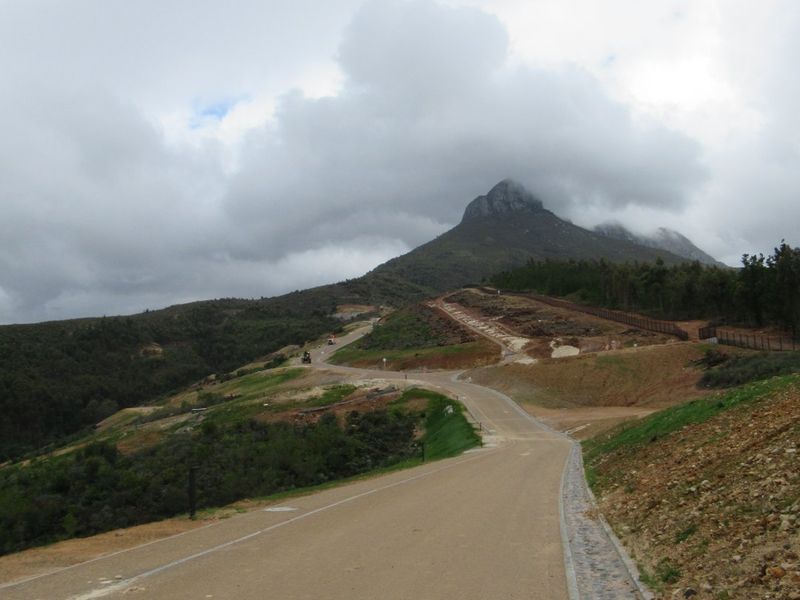 A view of a paved access road winding through a residential development with mountain views and established perimeter fencing.