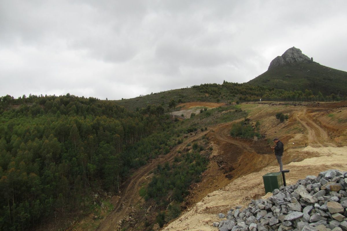 A wide-angle view of a sloped, undeveloped residential plot featuring natural vegetation, dirt access paths, and a prominent mountain backdrop.
