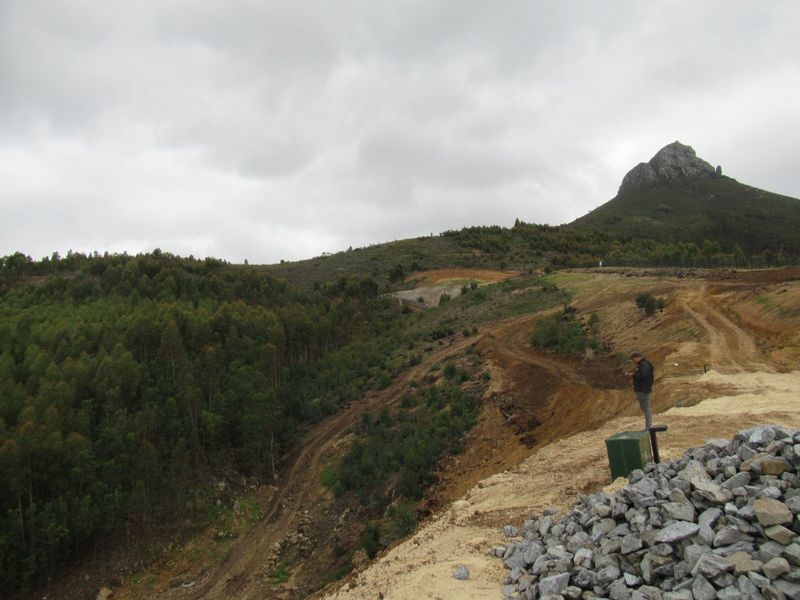 A wide-angle view of a sloped, undeveloped residential plot featuring natural vegetation, dirt access paths, and a prominent mountain backdrop.