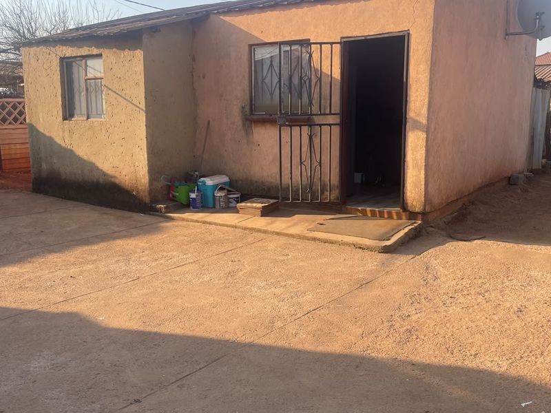 Rear view of a detached dwelling featuring a concrete apron, metal security gate, and weathered exterior plaster walls.