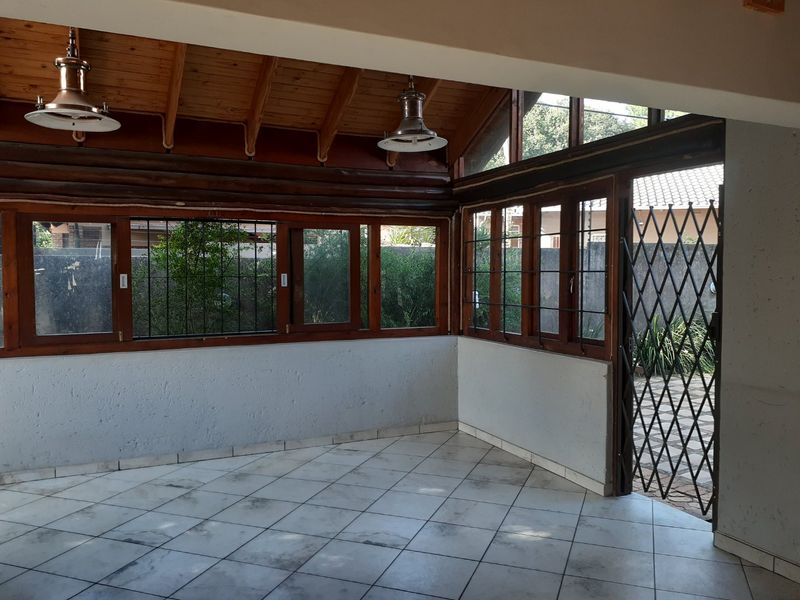 An empty covered patio area featuring exposed timber beam ceilings, tiled flooring, and large timber-framed windows with security gates leading to the garden.