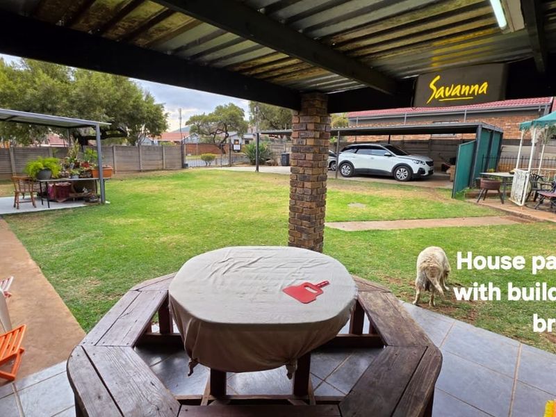 A covered outdoor patio area featuring tiled flooring, a brick support pillar, and a built-in braai, overlooking a spacious garden.