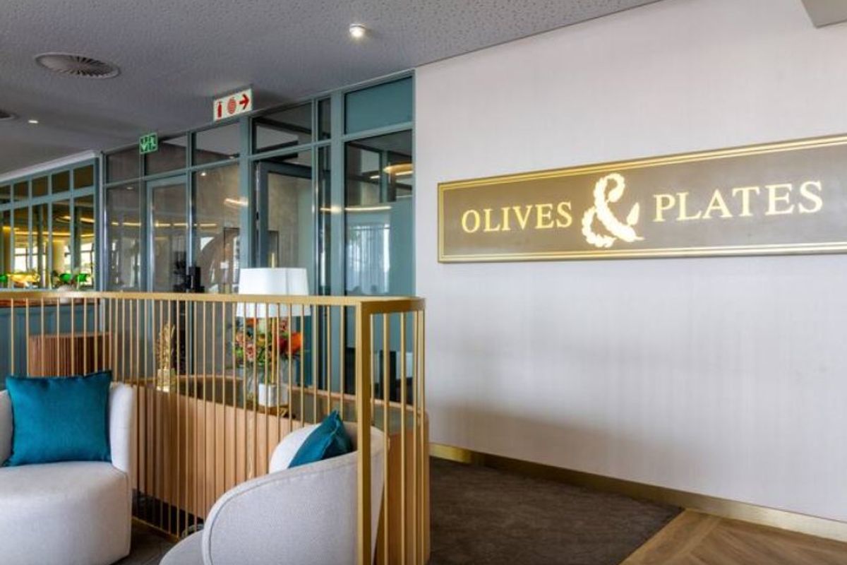 A modern commercial interior space featuring a gold metal balustrade, glass partitioning, and elegant herringbone flooring, currently branded as Olives & Plates.