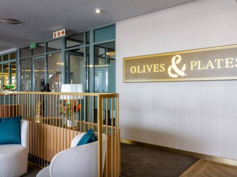 A modern commercial interior space featuring a gold metal balustrade, glass partitioning, and elegant herringbone flooring, currently branded as Olives & Plates.
