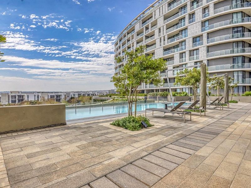 A modern outdoor pool area featuring a paved terrace, glass balustrades, and landscaped planters, set against a contemporary apartment building.