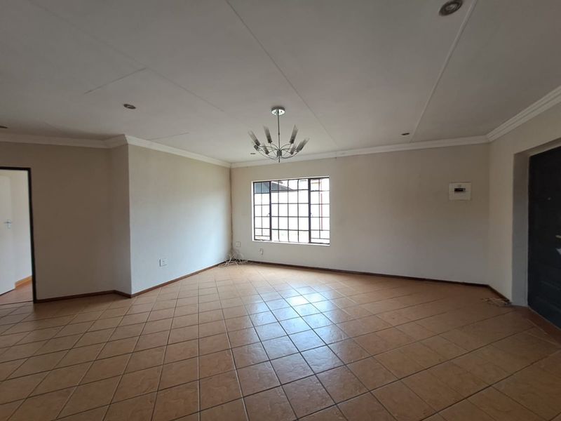 A spacious, empty living area featuring neutral walls, tiled flooring, a decorative chandelier, and a large grid-pattern window providing natural light.