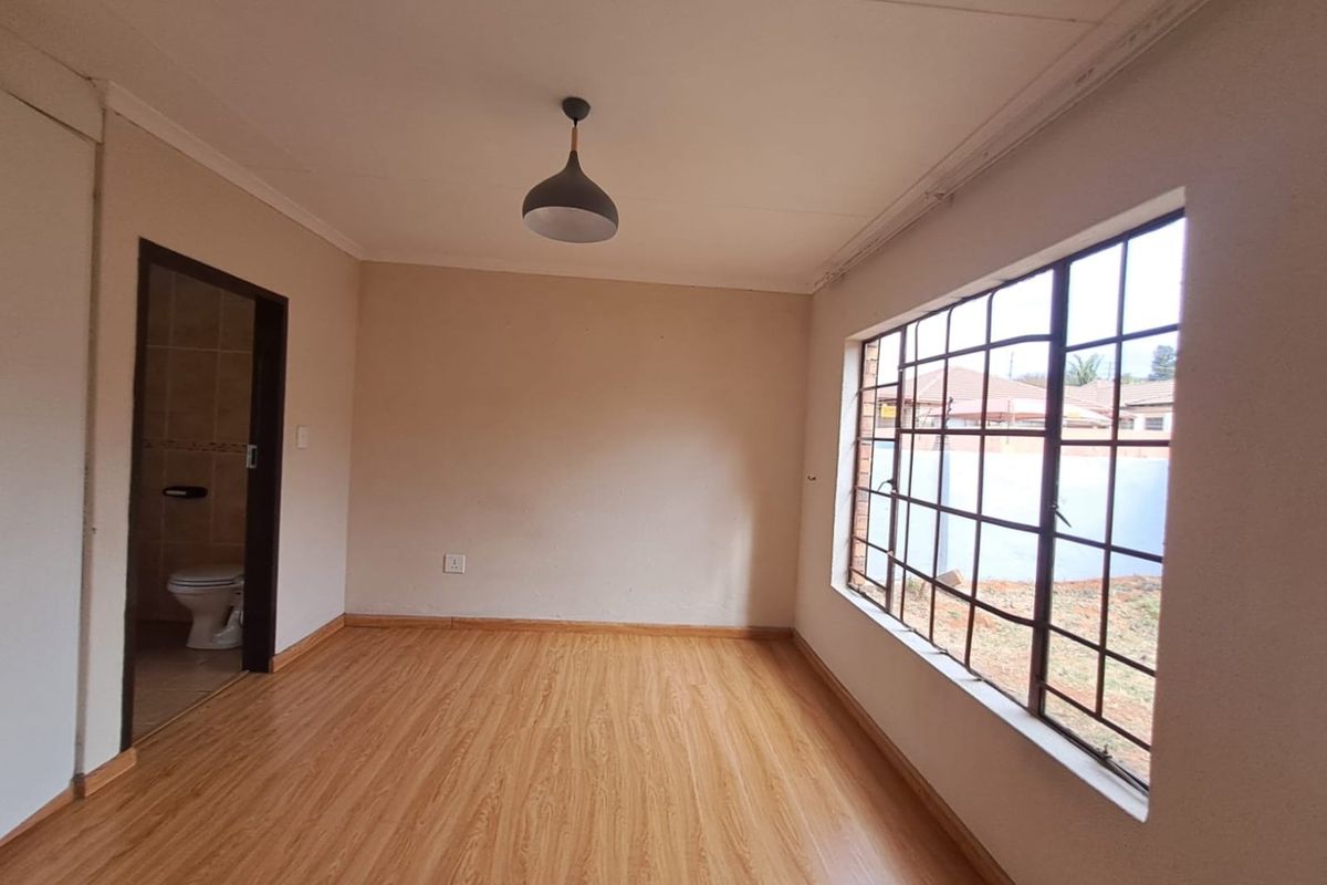 A bright, empty bedroom featuring laminate flooring, a pendant light, and an en-suite bathroom entrance, with metal security bars on the window.