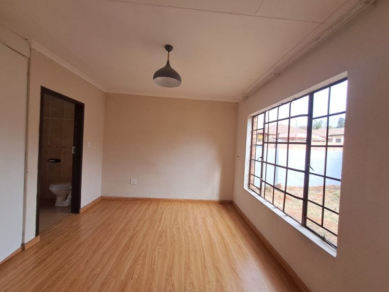 A bright, empty bedroom featuring laminate flooring, a pendant light, and an en-suite bathroom entrance, with metal security bars on the window.