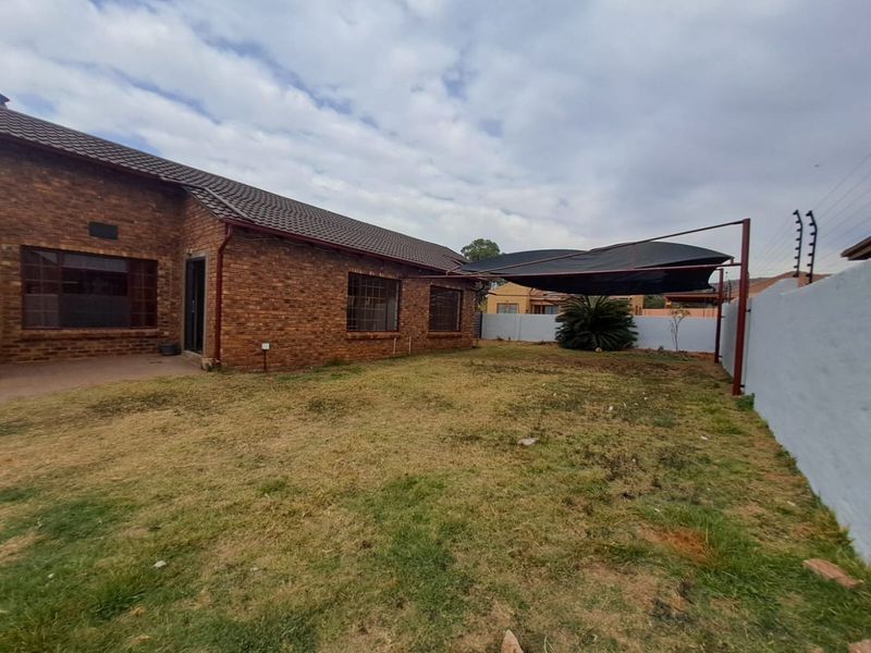 Rear view of a face brick house featuring a concrete patio, a shade cloth carport, and a perimeter wall with electric fencing.