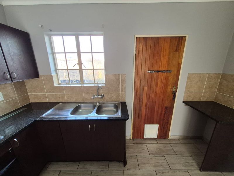 A functional kitchen area featuring dark wood cabinetry, granite countertops, a double bowl stainless steel sink, and tiled backsplash with a wooden exterior door.