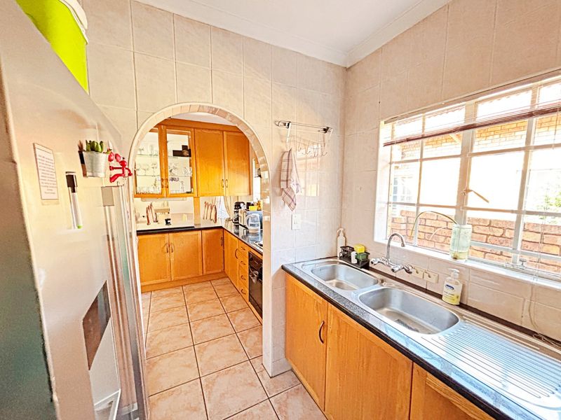 A functional kitchen featuring wooden cabinetry, a double stainless steel sink, tiled splashbacks, and an arched opening leading to an additional prep area.