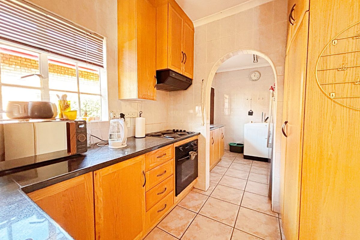 A functional kitchen featuring wooden cabinetry, built-in oven and hob, tiled splashbacks, and an arched transition to a secondary utility or laundry area.